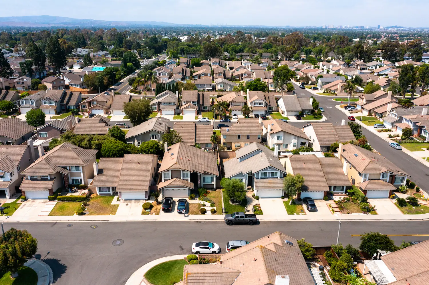 Aerial view of a suburban neighborhood with rows of similar houses, each with a driveway and cars. Streets are wide and lined with green trees. Calm and orderly setting.